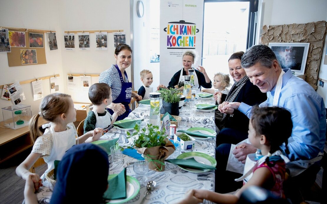 Stiftungsgründerin Sarah Wiener sitzt mit Kindern in einer Kita am gedeckten Tisch mit Nordrhein-Westfalens Verbraucherschutz- und Landwirtschaftsministerin Ursula Heinen-Esser und Heiner Beckmann. 