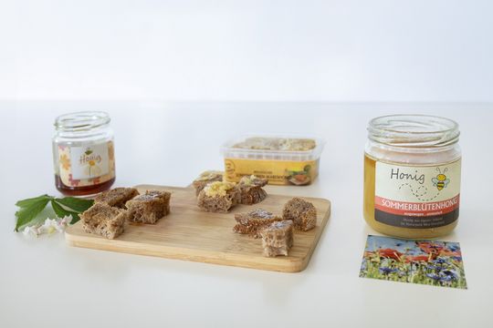 Auf einer weißen Fläche stehen drei Sorten Honig. Mittig ist ein Holzbrett platziert auf dem kleine Brothäppchen mit Honig zum probieren liegen.